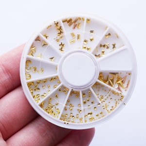 Op de afbeelding: Een wit plastic wiel met 12 vakken gevuld met goudkleurige nagelkunstversieringen. Het wiel is verdeeld in secties met verschillende vormen en maten versieringen, waaronder sterren, halve manen en kleine cirkels.