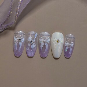 K&ouml;nnte beinhalten: Ein Satz von f&uuml;nf k&uuml;nstlichen N&auml;geln mit einem hellvioletten und wei&szlig;en Design. Jeder Nagel zeigt eine wei&szlig;e Schleife mit einem Strass- oder Perlenmittelpunkt. Die N&auml;gel haben ein gl&auml;nzendes Finish und sind auf einer neutralen Oberfl&auml;che angeordnet.