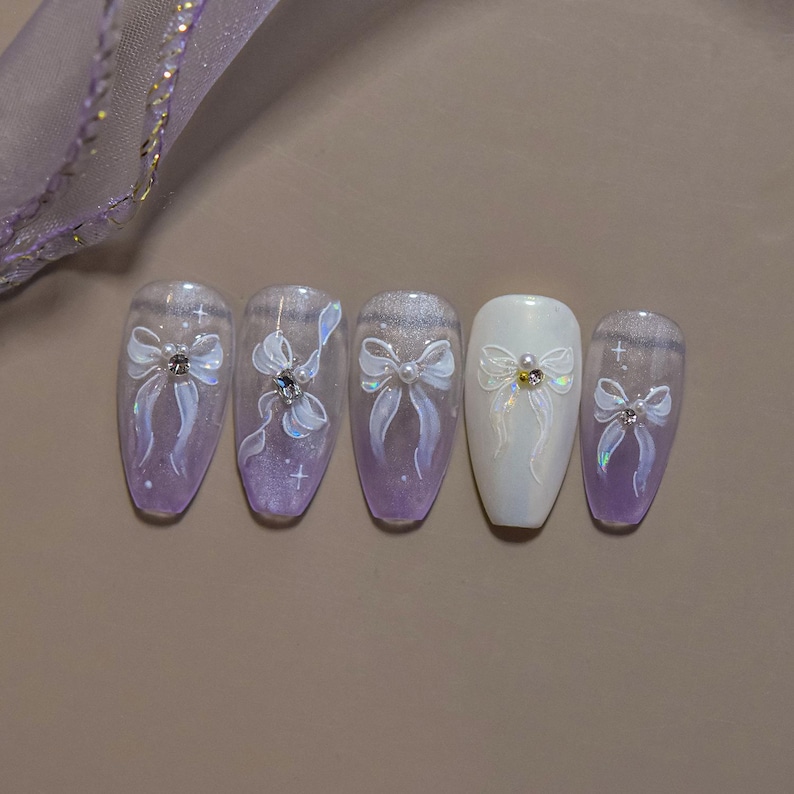 K&ouml;nnte beinhalten: Ein Satz von f&uuml;nf k&uuml;nstlichen N&auml;geln mit einem hellvioletten und wei&szlig;en Design. Jeder Nagel zeigt eine wei&szlig;e Schleife mit einem Strass- oder Perlenmittelpunkt. Die N&auml;gel haben ein gl&auml;nzendes Finish und sind auf einer neutralen Oberfl&auml;che angeordnet.