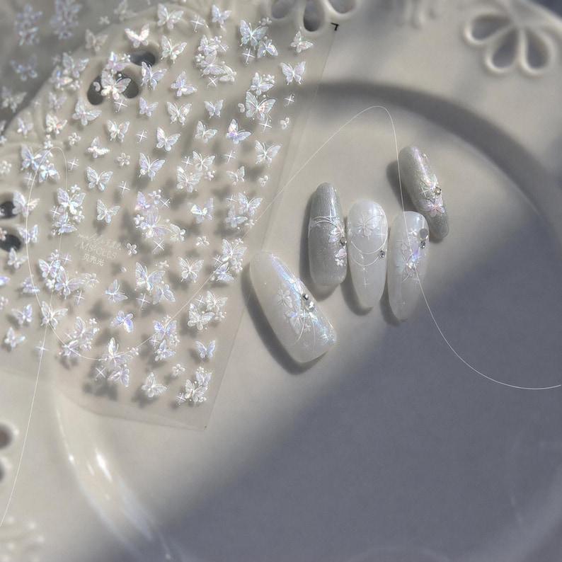 K&ouml;nnte beinhalten: Ein Bogen mit schillernden Schmetterlings-Nagelkunst-Aufklebern neben einem Satz manik&uuml;rter N&auml;gel. Die N&auml;gel sind wei&szlig; und silber lackiert und mit zarten Schmetterlings- und Blumendesigns versehen. Einige N&auml;gel haben kleine, funkelnde Verzierungen.