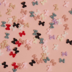 Puede incluir: Una colección de pequeños adornos en forma de lazo en varios colores, incluyendo negro, rojo, blanco, rosa y tonos pastel de azul, verde y morado. Los lazos están esparcidos sobre una superficie rosa claro.