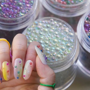 Könnte beinhalten: Nahaufnahme einer Hand mit mehreren ovalen Nägeln, die mit bunten, schillernden Perlen verziert sind. Die Nägel sind von vier kleinen Gläsern umgeben, die mit schillernden Perlen gefüllt sind.