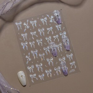 K&ouml;nnte beinhalten: Transparente Nagelkunst-Aufkleber mit schimmernden Schleifen und kleinen Kreuzen. Mehrere N&auml;gel sind mit lila und wei&szlig;en Designs verziert. Die Aufkleber befinden sich auf einem transparenten Bogen, mit einem kleinen Textetikett unten rechts.