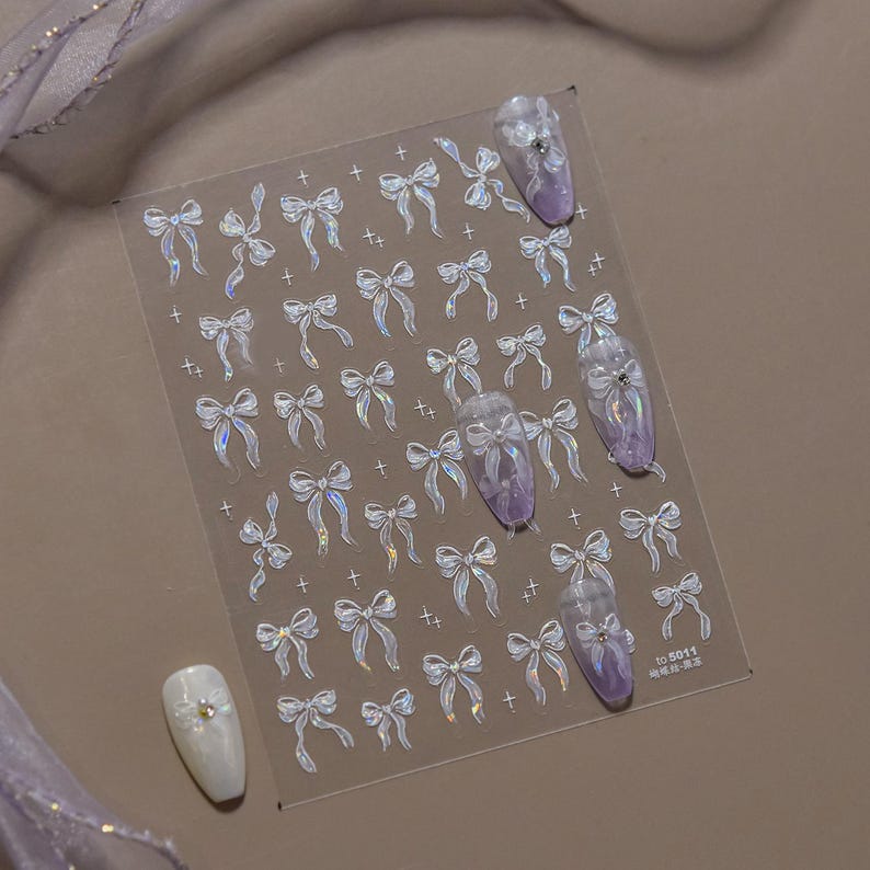 K&ouml;nnte beinhalten: Transparente Nagelkunst-Aufkleber mit schimmernden Schleifen und kleinen Kreuzen. Mehrere N&auml;gel sind mit lila und wei&szlig;en Designs verziert. Die Aufkleber befinden sich auf einem transparenten Bogen, mit einem kleinen Textetikett unten rechts.