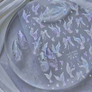 Può includere: Un foglio di adesivi per nail art a tema sirena. Gli adesivi presentano disegni traslucidi e iridescenti di code di sirena, fiocchi e altri elementi acquatici in tonalità viola, blu e bianco. Alcune unghie sono decorate con gli adesivi.