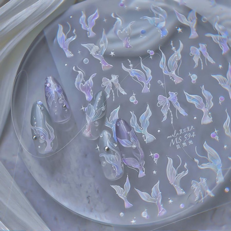 Peut inclure: Planche de stickers pour nail art sur le th&egrave;me des sir&egrave;nes. Les autocollants pr&eacute;sentent des motifs translucides et iris&eacute;s de queues de sir&egrave;nes, de n&oelig;uds et d'&eacute;l&eacute;ments aquatiques dans des tons violets, bleus et blancs. Des ongles sont d&eacute;cor&eacute;s avec les autocollants.