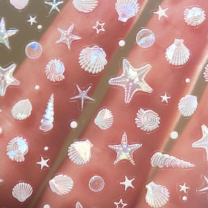 Peut inclure: Feuille transparente d'autocollants iris&eacute;s avec diverses formes sur le th&egrave;me de la mer. Les autocollants comprennent des &eacute;toiles de mer, des coquillages, des coquilles en spirale et de petites &eacute;toiles. Les autocollants ont une finition chatoyante et nacr&eacute;e.
