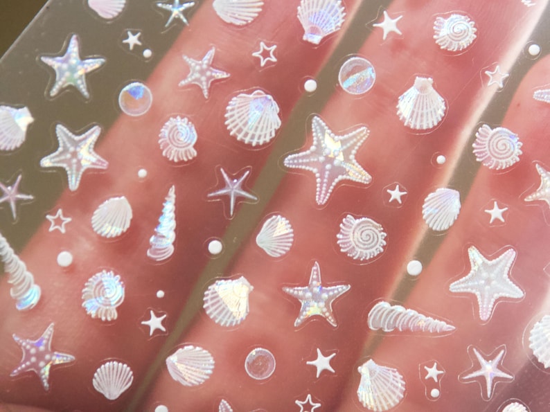 Peut inclure: Feuille transparente d'autocollants iris&eacute;s avec diverses formes sur le th&egrave;me de la mer. Les autocollants comprennent des &eacute;toiles de mer, des coquillages, des coquilles en spirale et de petites &eacute;toiles. Les autocollants ont une finition chatoyante et nacr&eacute;e.