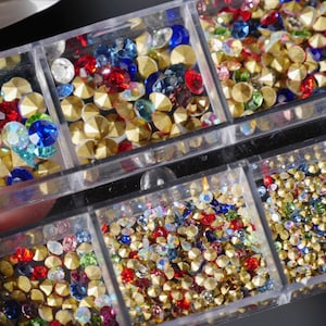 以下が含まれることがあります： さまざまな色のラインストーンが詰められた透明なプラスチック製のオーガナイザー。 ラインストーンは、ゴールド、レッド、ブルー、イリデセントカラーです。 コンパートメントには、さまざまなサイズと形状の装飾石が詰められています。