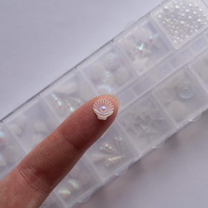 May include: A small, iridescent seashell-shaped nail art decoration is held by a finger. The seashell is white with a pearlescent sheen and a small, pink center. A clear plastic organizer filled with various nail art supplies is in the background.