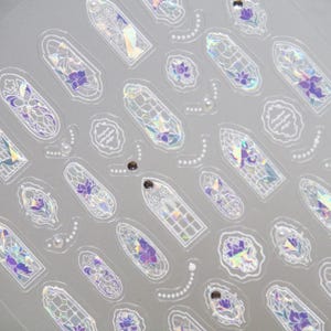 May include: A sheet of clear stickers featuring a repeating pattern of stained-glass window designs. The designs include purple floral accents, iridescent details, and decorative white outlines. Small pearl-like embellishments are also present.