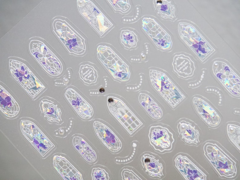 May include: A sheet of clear stickers featuring a repeating pattern of stained-glass window designs. The designs include purple floral accents, iridescent details, and decorative white outlines. Small pearl-like embellishments are also present.
