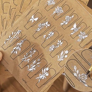Puede incluir: Hojas de pegatinas de arte de uñas con diseños florales blancos sobre un fondo marrón. Cada pegatina tiene un borde negro, con diseños que incluyen rosas, hojas y otros elementos botánicos. Las hojas están dispuestas en forma de abanico.