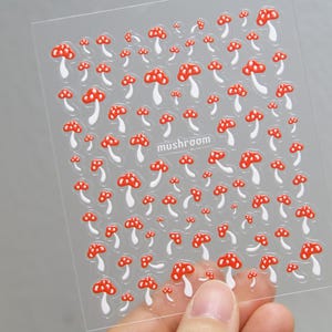 May include: Clear sheet of stickers featuring a pattern of red and white mushrooms. The word "mushroom" is printed in a small font. The stickers are held by a person's hand.
