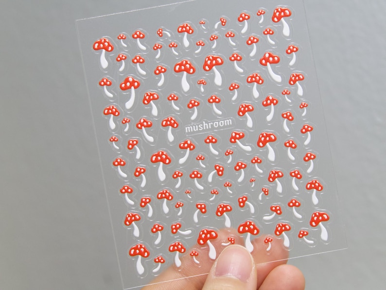 May include: Clear sheet of stickers featuring a pattern of red and white mushrooms. The word "mushroom" is printed in a small font. The stickers are held by a person's hand.