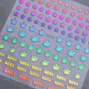 May include: Sheet of nail art stickers featuring cannabis leaf designs in a variety of colors, including green, blue, purple, and orange. The word "DOPE" is printed in a dripping font at the bottom of the sheet.