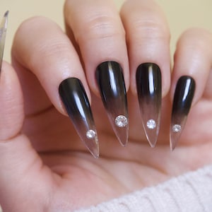 May include: Close-up of a hand with long, stiletto-shaped nails. The nails are painted with a black to clear ombre effect and have a small clear rhinestone on each nail.