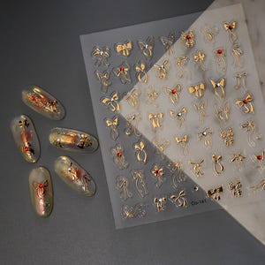 Puede incluir: Una l&aacute;mina de pegatinas de u&ntilde;as con lazos dorados con detalles rojos y transparentes. Tambi&eacute;n se ven varios dise&ntilde;os de u&ntilde;as con lazos dorados y detalles rojos. La l&aacute;mina tiene el texto "Co-141" impreso.