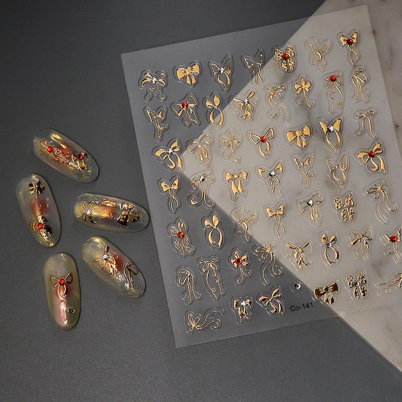 Puede incluir: Una l&aacute;mina de pegatinas de u&ntilde;as con lazos dorados con detalles rojos y transparentes. Tambi&eacute;n se ven varios dise&ntilde;os de u&ntilde;as con lazos dorados y detalles rojos. La l&aacute;mina tiene el texto "Co-141" impreso.