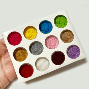 Pode incluir: Uma caixa retangular branca com doze aberturas circulares, cada uma preenchida com uma bola de feltro colorida. As cores incluem vermelho, amarelo, azul, verde, castanho, rosa, cinzento, branco e roxo.