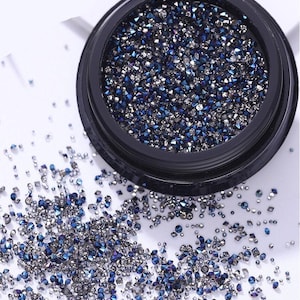 May include: A small black container filled with iridescent blue and silver rhinestones. The container is open and some rhinestones have spilled out onto a white surface.