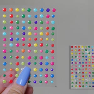 Puede incluir: Dos hojas de pegatinas redondas en 3D de colores. Las pegatinas están dispuestas en forma de cuadrícula y son de varios colores, incluidos rojo, naranja, amarillo, verde, azul, morado y rosa.