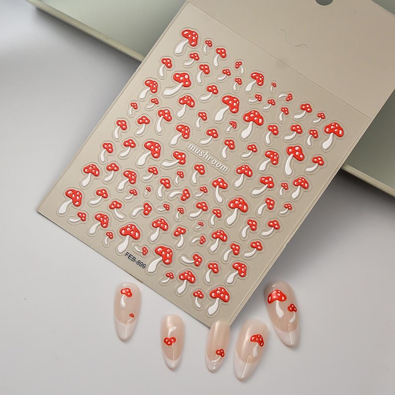 May include: A sheet of nail art stickers featuring a repeating pattern of red and white mushroom designs. The word "mushroom" is printed on the sheet. Several manicured nails with mushroom designs are also visible.
