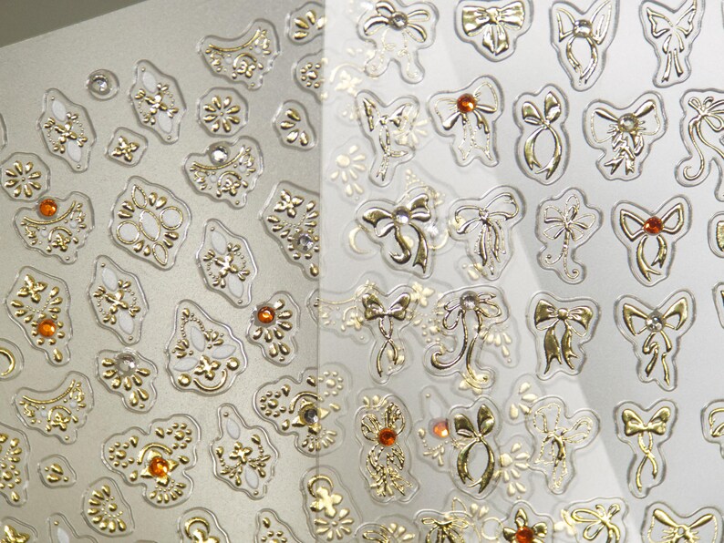 Puede incluir: Hoja de pegatinas decorativas con adornos dorados y transparentes. Los dise&ntilde;os incluyen formas ornamentadas, lazos y motivos florales. Algunas pegatinas tienen peque&ntilde;as gemas de color naranja. Las pegatinas est&aacute;n sobre un fondo gris claro.