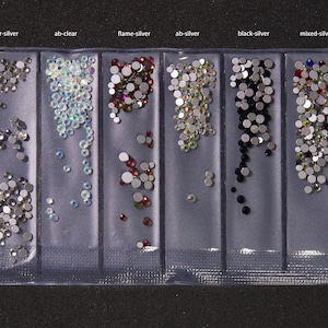 Puede incluir: Un recipiente de plástico transparente dividido en seis secciones, cada una llena de pequeños pedrería redondos. Las secciones están etiquetadas: clear-silver, ab-clear, flame-silver, ab-silver, black-silver y mixed-silver. La pedrería varía en color y acabado.