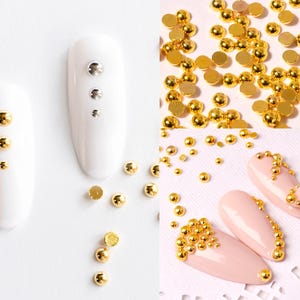 May include: Gold nail embellishments in various sizes, including small round beads and larger round beads with a raised center. The beads are shown on a white background and on a fingernail.