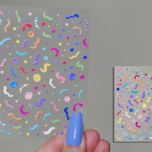 May include: A sheet of colorful nail stickers with a variety of shapes and sizes. The stickers are on a clear background and feature a mix of colors, including pink, blue, green, yellow, and orange.
