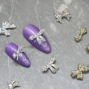 May include: A set of silver and gold nail charms in the shape of bows. The charms are decorated with rhinestones and are displayed on purple nail tips.