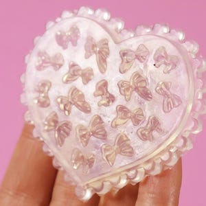 Puede incluir: Un molde en forma de corazón con borde festoneado, lleno de pequeñas formas de lazo. El molde es de color translúcido e iridiscente, sobre un fondo rosa. Los lazos están espaciados uniformemente dentro del corazón.
