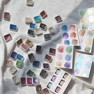 Könnte beinhalten: Eine Sammlung von metallischen Aquarellfarben in kleinen quadratischen Schalen. Die Schalen sind in einem zufälligen Muster auf einer weißen Stoffoberfläche angeordnet. Die Farben sind in verschiedenen Schattierungen von Gold, Silber, Kupfer und Blau. Es gibt auch mehrere Farbmuster auf weißem Papier.