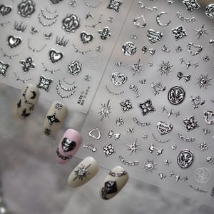May include: Silver nail art stickers with various designs, including hearts, stars, moons, and chains. The stickers are displayed with painted nails in different colors, showcasing the application of the nail art. The stickers are on a clear sheet.
