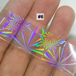 May include: A clear, holographic nail art strip with a repeating starburst pattern in shades of purple, green, and yellow. The strip is held in a hand and has the number "#6" printed on it. The nail art strip is designed for decorative purposes.