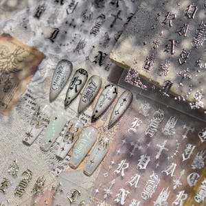 Könnte beinhalten: Eine Sammlung von Nagelkunstzubehör mit gotischen Designs. Das Bild zeigt verschiedene Nagelkunst-Elemente, darunter Nagelaufkleber mit Kreuzen, Buchstaben und verzierten Mustern, sowie mehrere dekorierte Kunstnägel. Die Farbpalette umfasst Silber, Schwarz und Weiß.