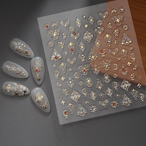 Puede incluir: Una l&aacute;mina de pegatinas para u&ntilde;as con adornos dorados y rojos con forma de joya. Las pegatinas presentan varios dise&ntilde;os ornamentados, incluyendo patrones florales y geom&eacute;tricos. Varias u&ntilde;as artificiales est&aacute;n decoradas con dise&ntilde;os similares.