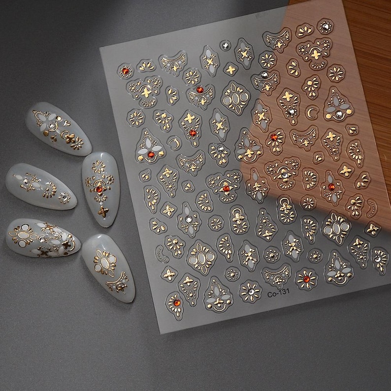 Puede incluir: Una l&aacute;mina de pegatinas para u&ntilde;as con adornos dorados y rojos con forma de joya. Las pegatinas presentan varios dise&ntilde;os ornamentados, incluyendo patrones florales y geom&eacute;tricos. Varias u&ntilde;as artificiales est&aacute;n decoradas con dise&ntilde;os similares.