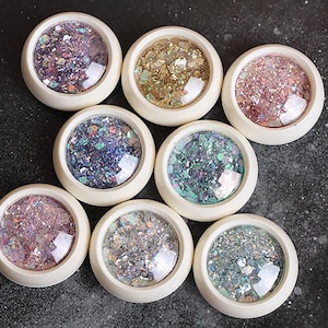 May include: Eight small round containers with clear lids filled with different colors of glitter. The containers are white with a silver rim.