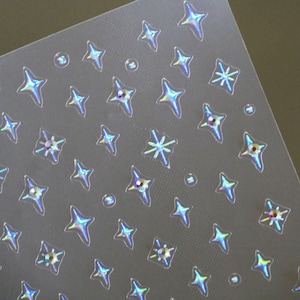 Puede incluir: Una hoja de pegatinas iridiscentes en forma de estrella y diamante. Las pegatinas están en una hoja de plástico transparente.