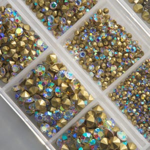 May include: A clear plastic organizer filled with various sizes of gold-colored, faceted studs and iridescent rhinestones. The studs have a metallic finish, and the rhinestones sparkle with multiple colors. Ideal for crafting and embellishments.