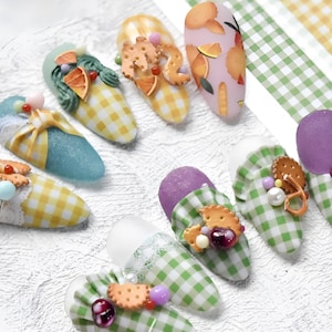 Puede incluir: Un conjunto de diez uñas postizas con varios diseños. Las uñas están decoradas con patrones de cuadros, frutas y galletas. Algunas uñas son moradas, azules y amarillas.