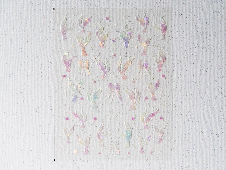 Peut inclure: Une feuille d'autocollants iris&eacute;s avec des motifs de queues de sir&egrave;ne et de n&oelig;uds. Les autocollants ont une finition nacr&eacute;e avec des nuances de rose, de violet et de bleu. De petits points roses sont dispers&eacute;s autour des motifs.