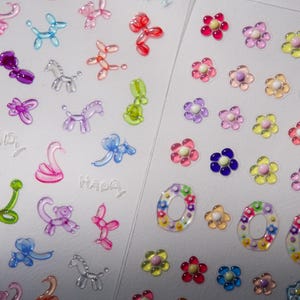 May include: Two sheets of colorful stickers. One sheet features a variety of balloon animal stickers, including a swan, a dog, a horse, and a unicorn. The other sheet features a variety of flower stickers in different colors, including pink, purple, yellow, and blue. Both sheets have the word "Happy" printed on them.