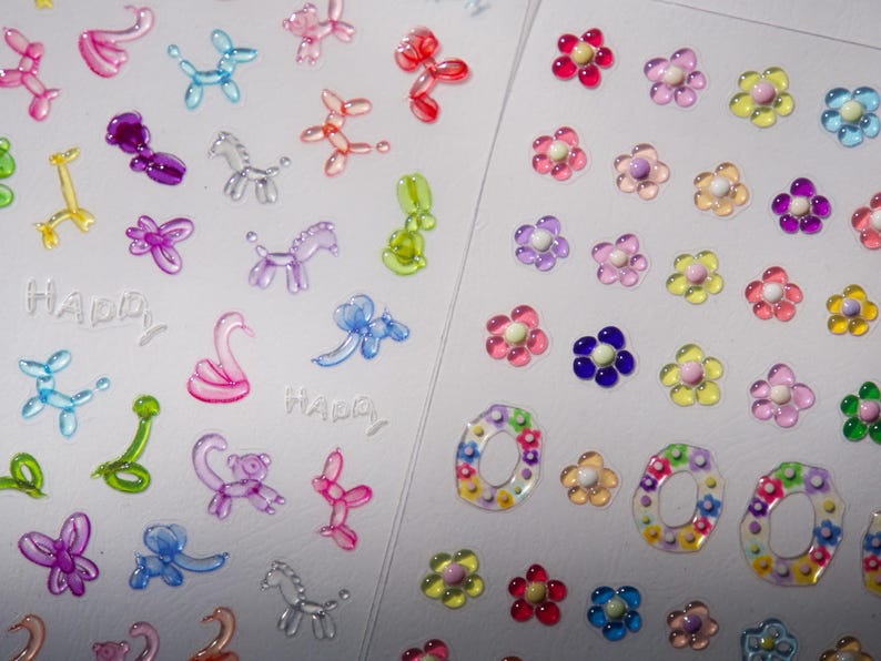 May include: Two sheets of colorful stickers. One sheet features a variety of balloon animal stickers, including a swan, a dog, a horse, and a unicorn. The other sheet features a variety of flower stickers in different colors, including pink, purple, yellow, and blue. Both sheets have the word "Happy" printed on them.