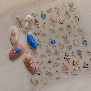 May include: A sheet of adhesive nail art stickers featuring gold metallic designs of moons, stars, lanterns, and ornate frames, alongside five decorated artificial fingernails displaying various celestial and magical motifs in blue, pink, and white.