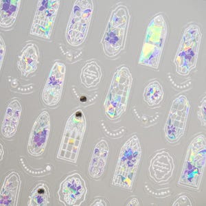 May include: Sheet of iridescent stickers featuring various shapes, including arched windows, floral designs, and decorative frames. The stickers have a holographic effect with purple and green hues, and some include pearl accents.