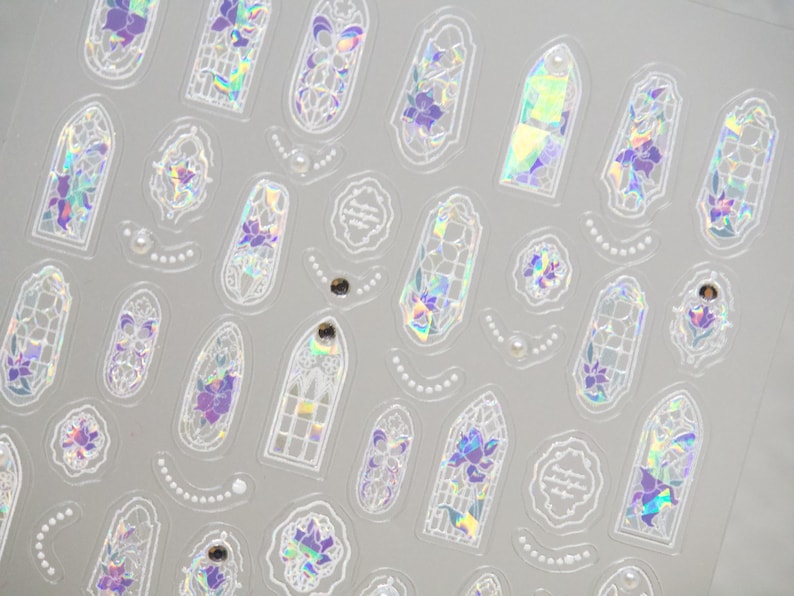 May include: Sheet of iridescent stickers featuring various shapes, including arched windows, floral designs, and decorative frames. The stickers have a holographic effect with purple and green hues, and some include pearl accents.