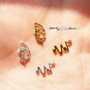 May include: Four gold and silver nail art charms. Two charms are in the shape of a heart beat line, and two charms are in the shape of a face profile.